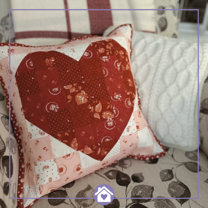 Scrappy heart pillow cover made with red and pink quilting cottons, styled on a floral chair with a plaid throw and cable-knit pillow for cozy Valentine’s Day décor.